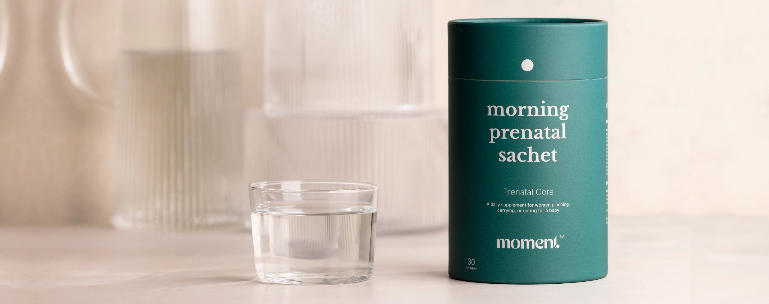 A teal cylindrical container labeled 'morning prenatal sachet' stands on a light surface beside a small clear glass cup with liquid, against a softly blurred warm neutral background.