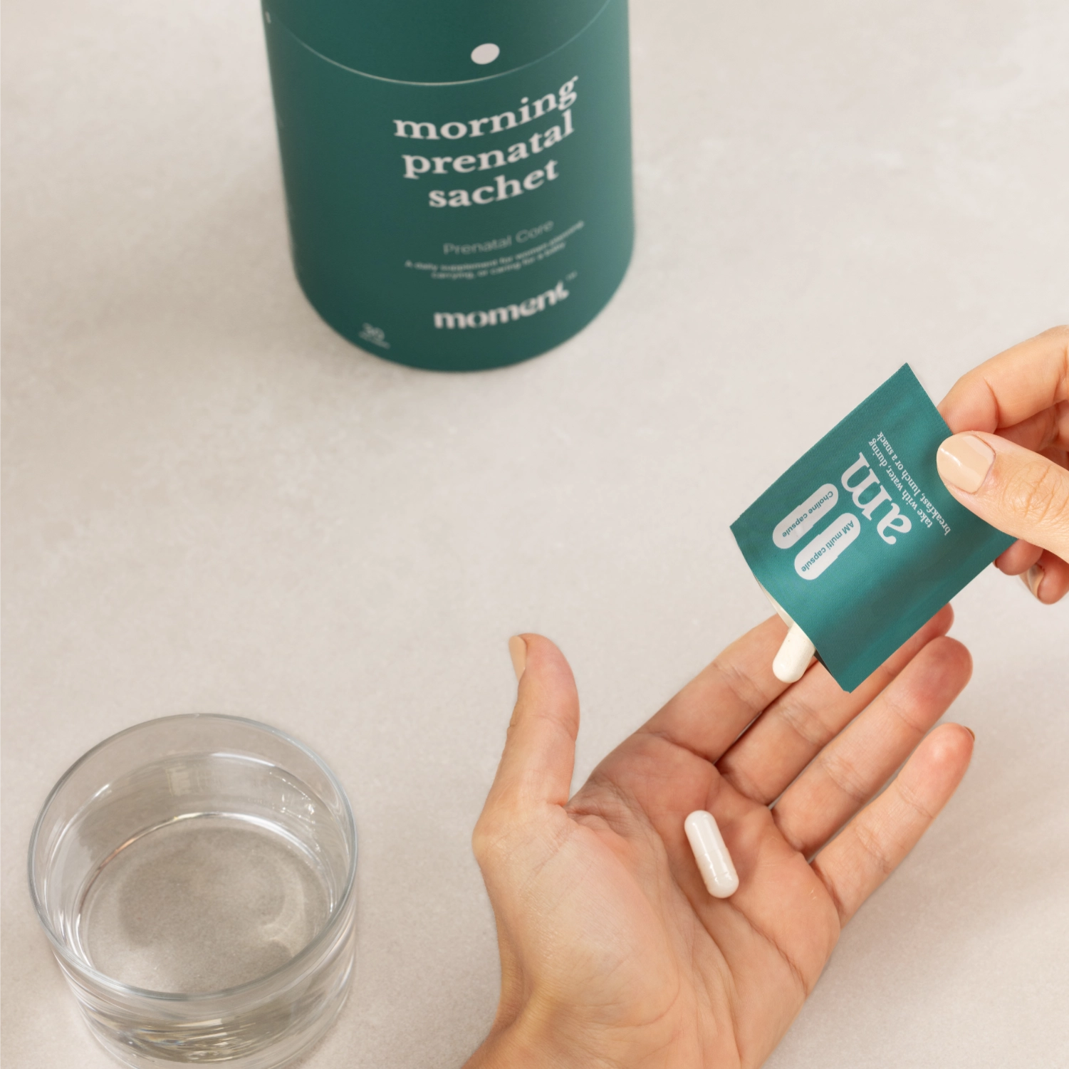A person holds a small teal sachet labeled 'am' over their open palm pouring vitamins out of the sachet into their hand, with a larger teal bottle labeled 'morning prenatal sachet' in the background and a glass of water in the foreground.