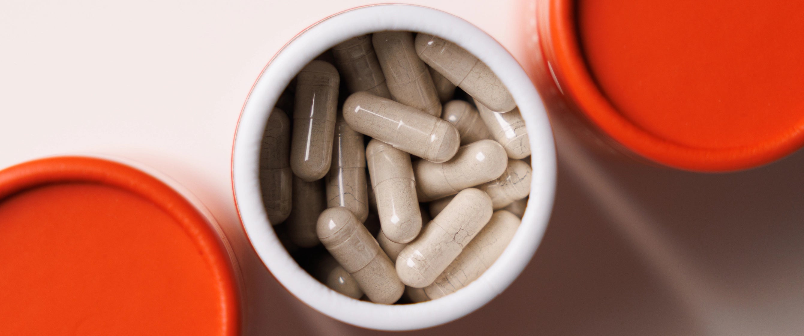 Top-down view of off-white capsules inside an open white pill bottle with an orange cap, with two more orange caps partially visible on the left and right.