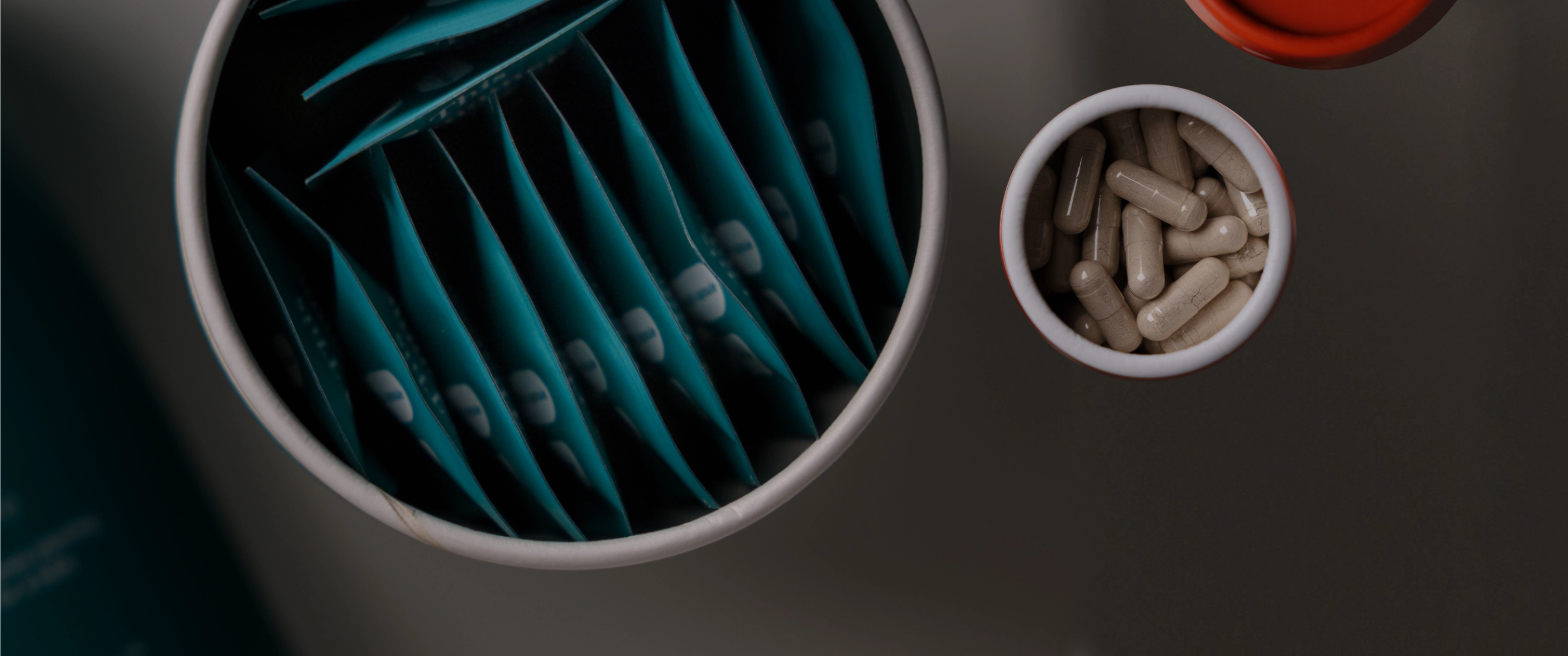 A top-down view of a supplement container with multiple teal-blue sachets inside and a smaller orange and white canister filled with beige capsules, set on a dark gray surface.
