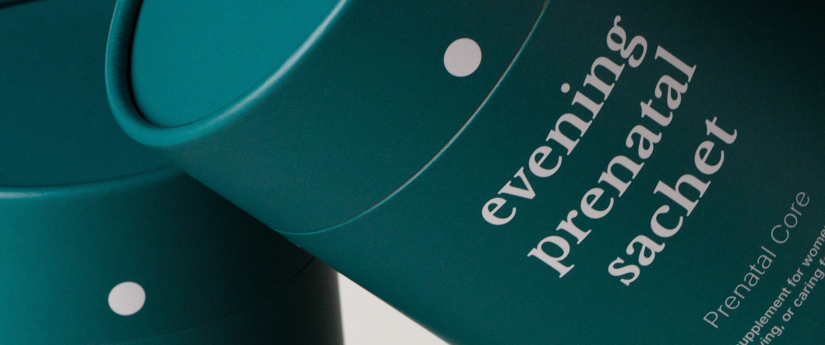 Close-up of teal cylindrical supplement packaging with white text reading 'evening prenatal sachet' and small white dots, shown on a light neutral background.